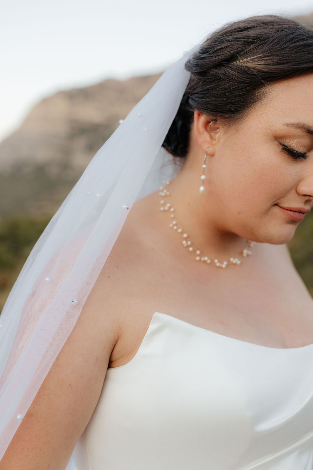 Bride in strapless white gown wearing pearl necklace and earrings with flowing veil against mountain backdrop.