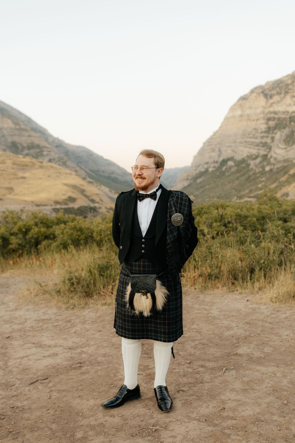 Groom in traditional Scottish kilt with sporran and black jacket standing before mountain landscape.