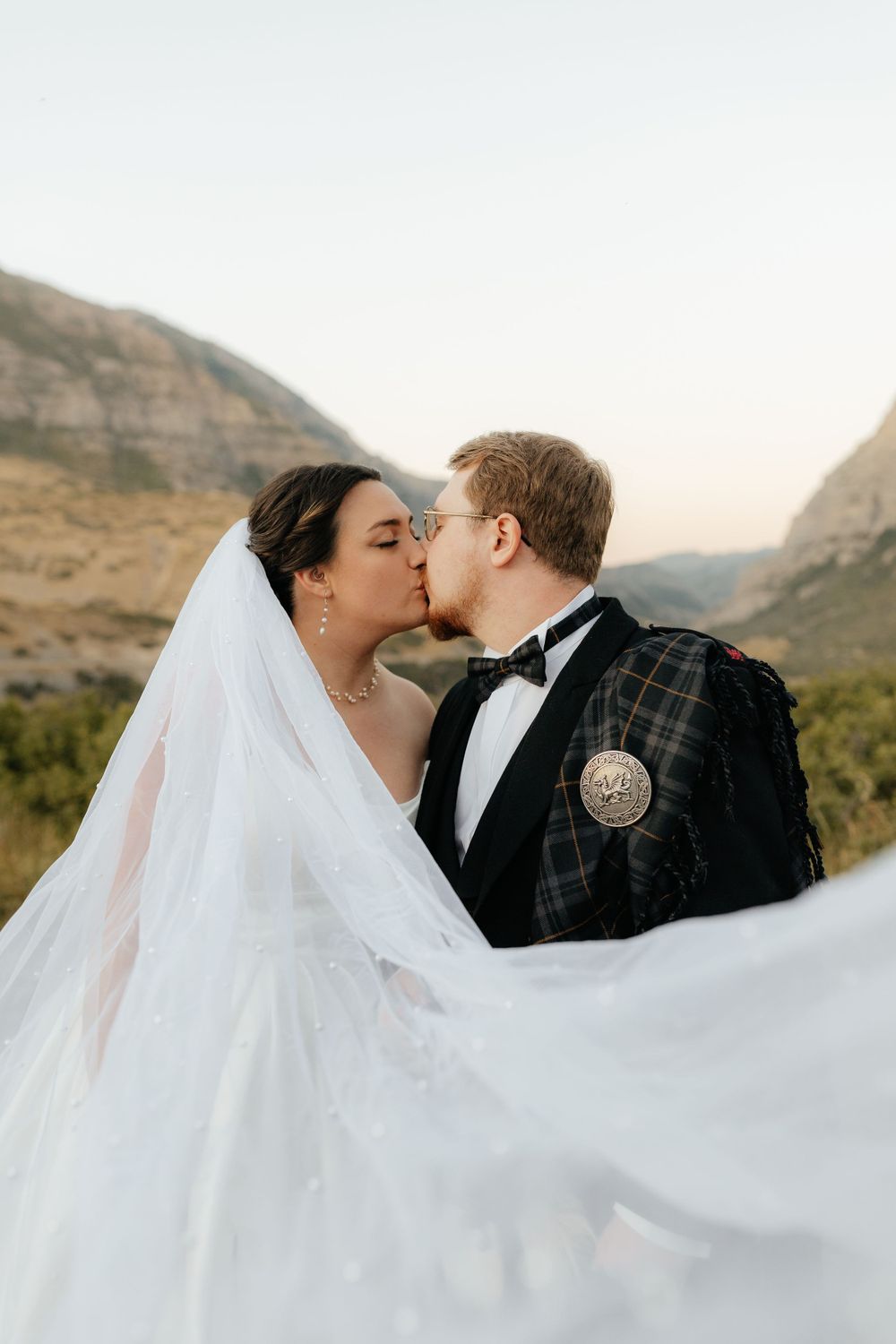 Bride and groom sharing a kiss with flowing white veil billowing in mountain setting.