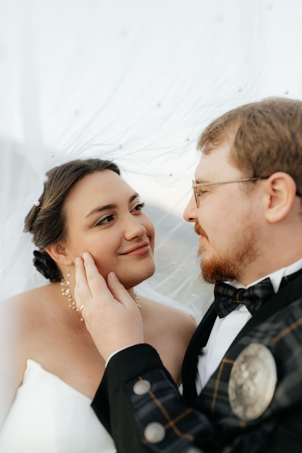 Groom tenderly cupping bride's face under flowing white veil in intimate wedding portrait.