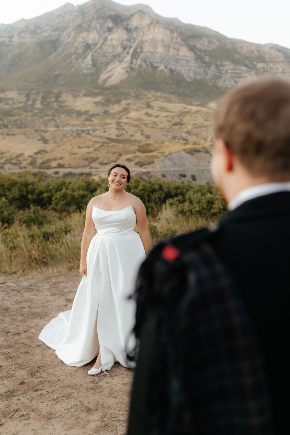 Smiling bride in strapless white gown stands before rugged mountain landscape with groom in foreground.