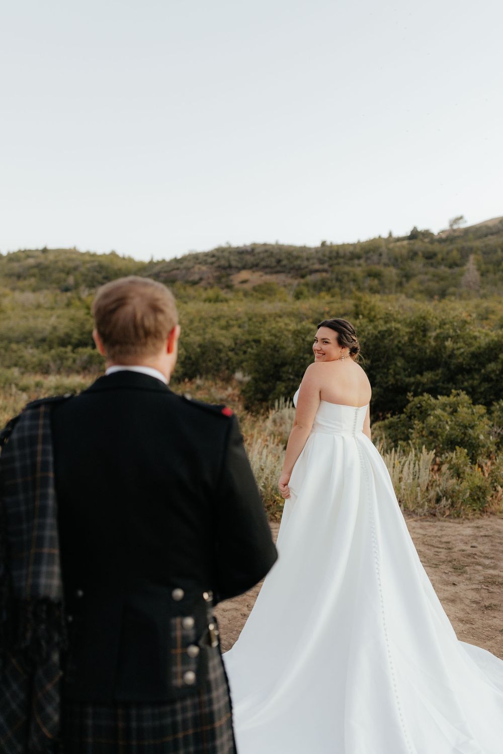 Bride in flowing white gown looks back smiling at groom in Scottish kilt on mountain trail.