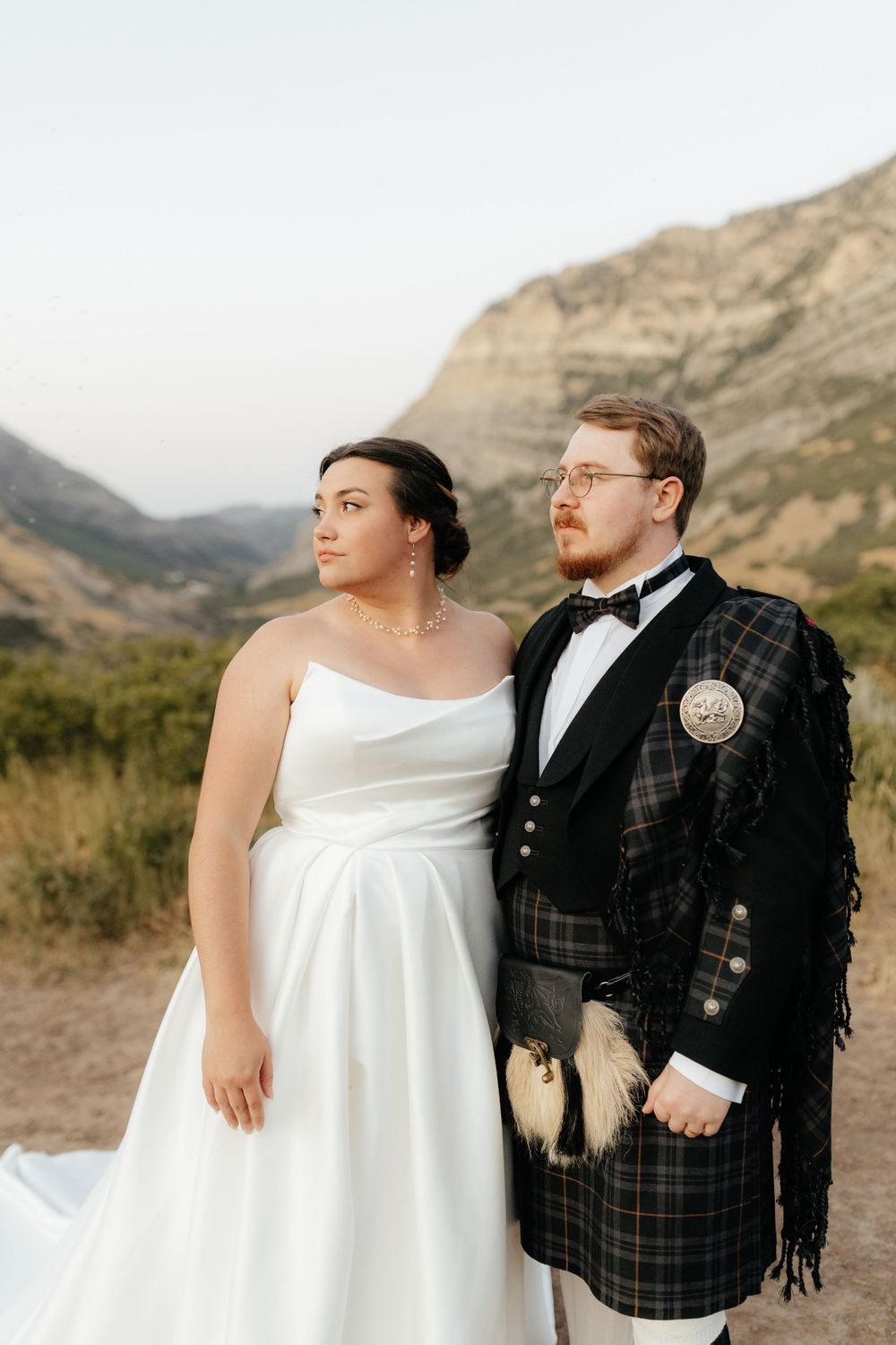 Bride and groom in Scottish kilt attire pose together gazing at stunning mountain scenery.