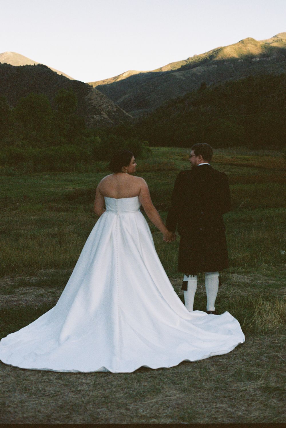 Bride and groom holding hands walking toward scenic mountain valley at golden hour sunset.