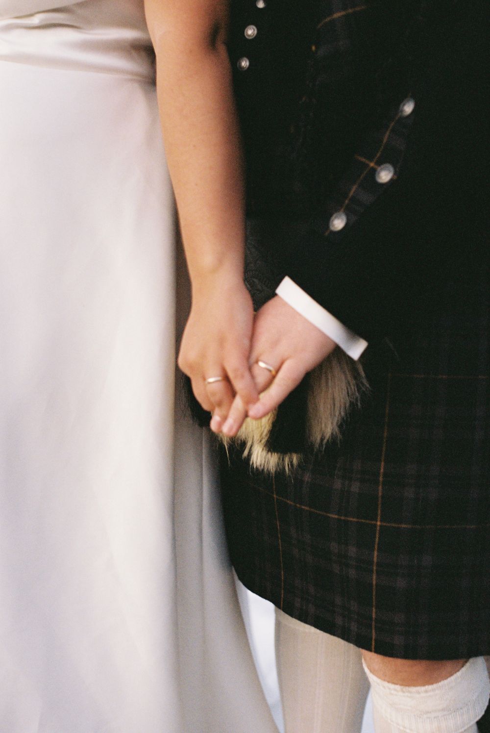 Close-up of bride and groom holding hands showing wedding rings beside Scottish kilt sporran.
