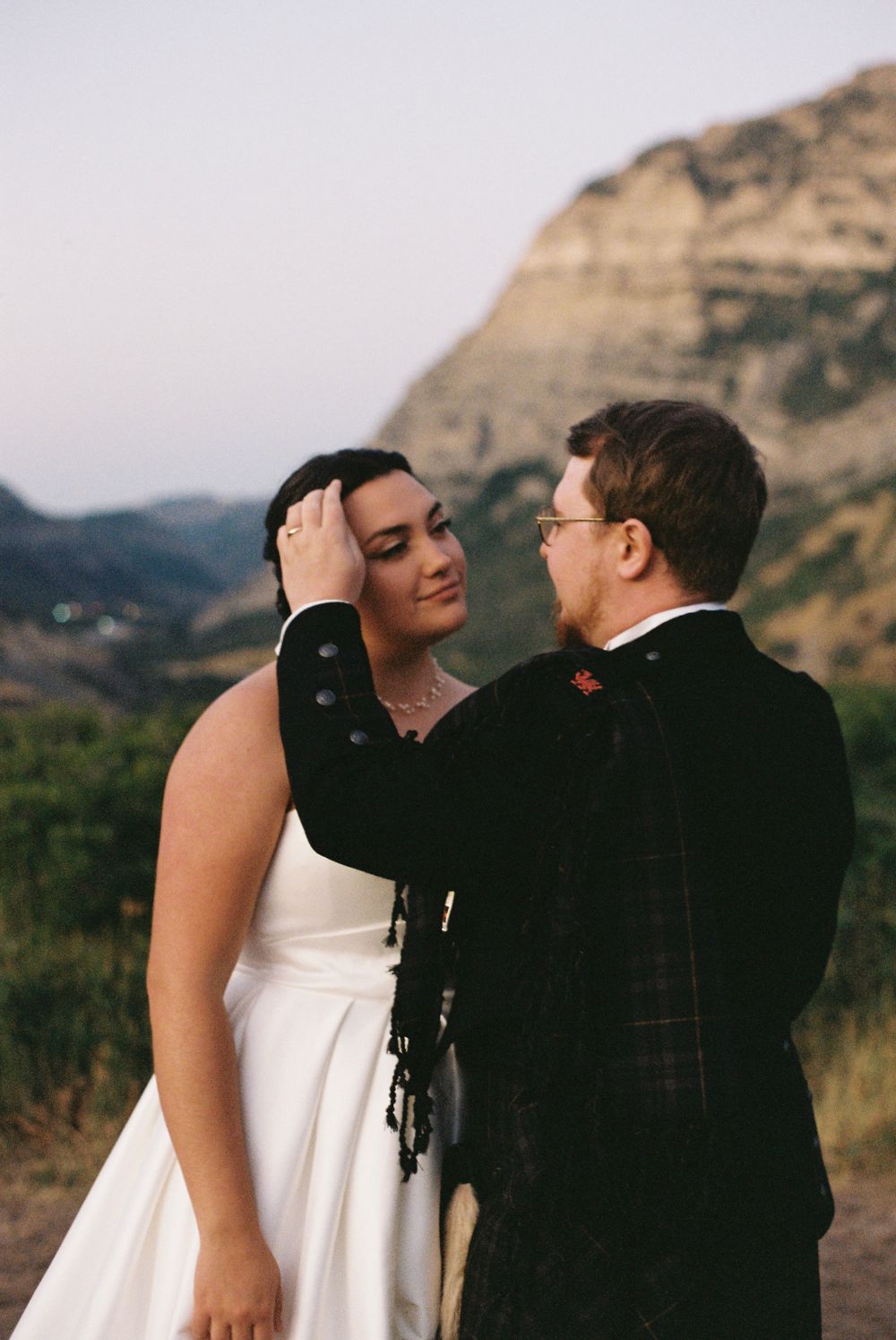 Groom in Scottish tartan kilt tenderly touches bride's face during romantic mountain wedding portrait at sunset.