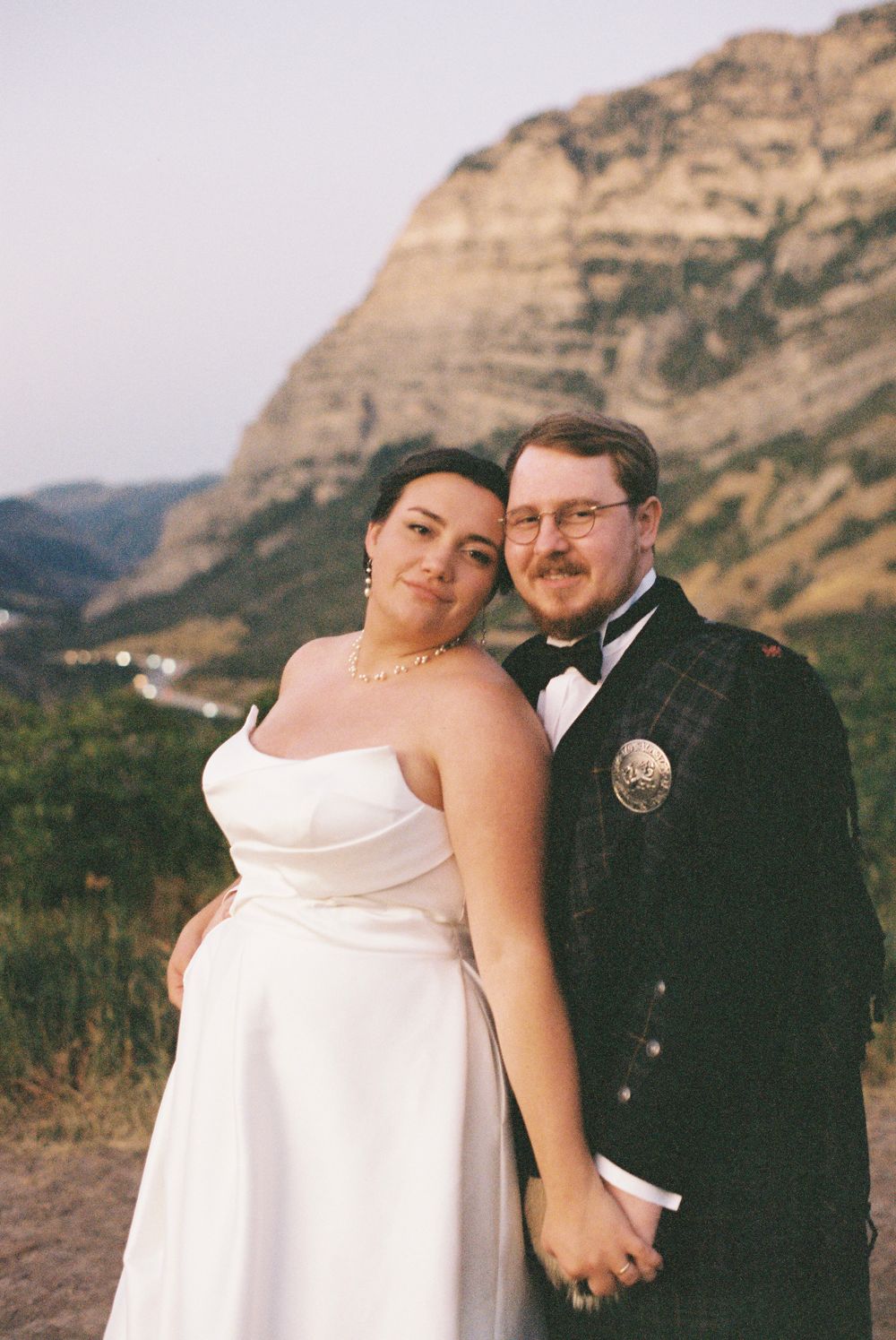 Bride and groom pose together in front of stunning mountain backdrop, groom wearing traditional Scottish tartan kilt.
