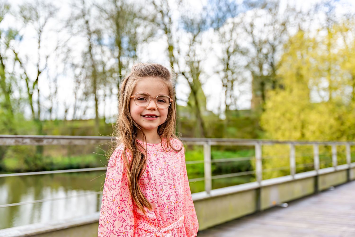A child in pink dress and glasses smiles while standing on a wooden boardwalk near water.