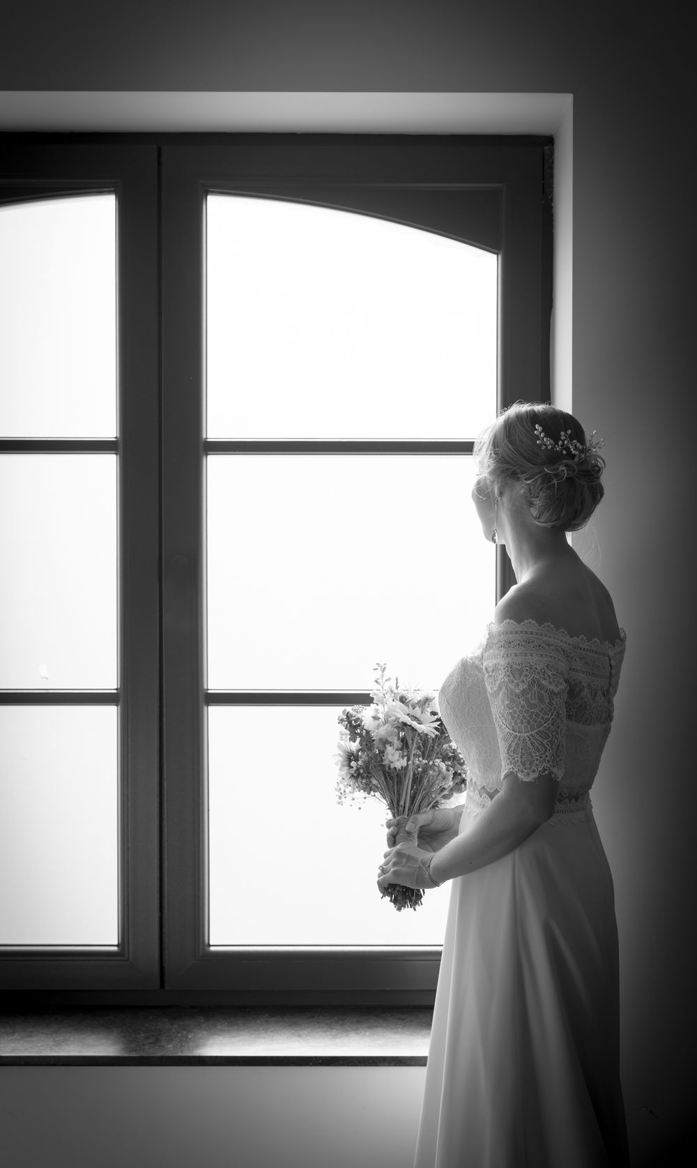 Silhouette of bride holding bouquet while standing by large window in dramatic lighting.