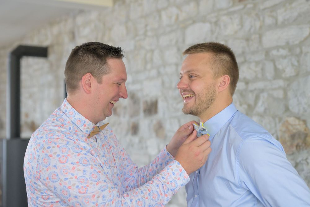 Groomsmen laughing together against stone wall backdrop during wedding celebrations.