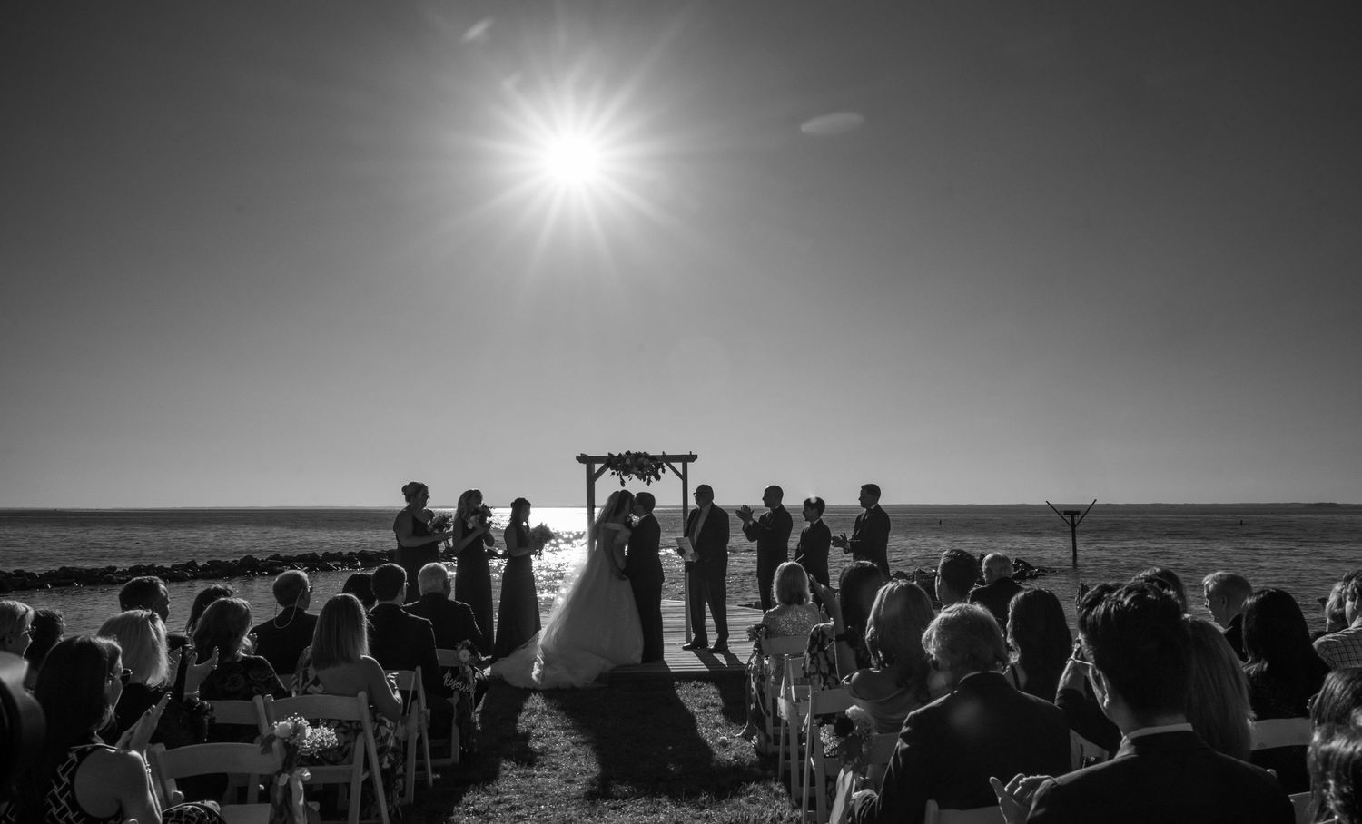 A black and white silhouette of a beach wedding ceremony with guests seated under the bright sun.