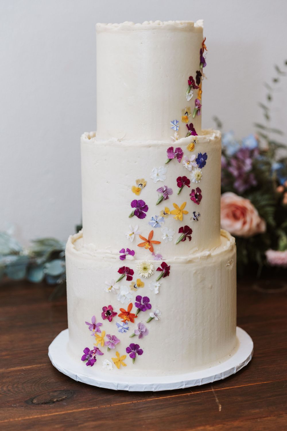 Three-tiered white wedding cake decorated with cascading colorful edible flowers and petals.