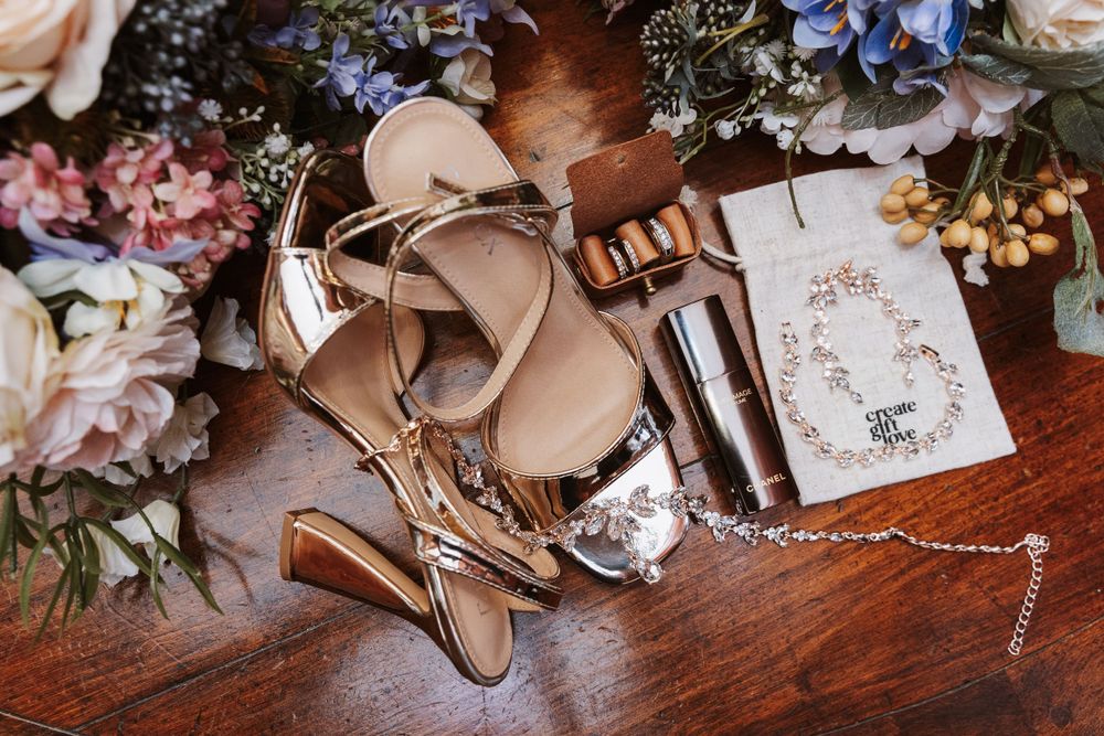 Wedding accessories including silver heels, jewelry, and floral arrangements on wooden surface.