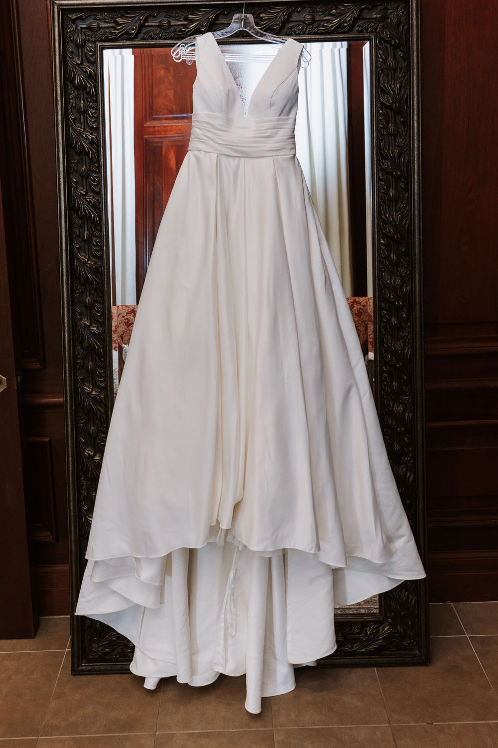 Classic white wedding gown with V-neckline hanging in ornate mirror frame.