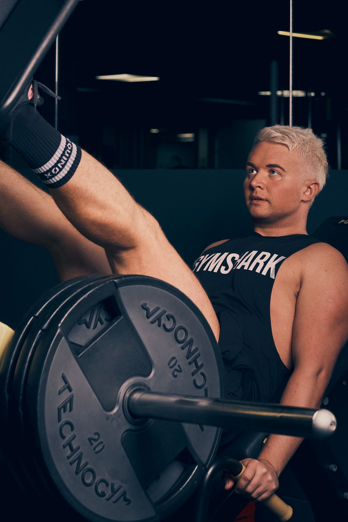 Man with blonde hair lifting weights in a gym, wearing a black Gymshark vest, focusing on form