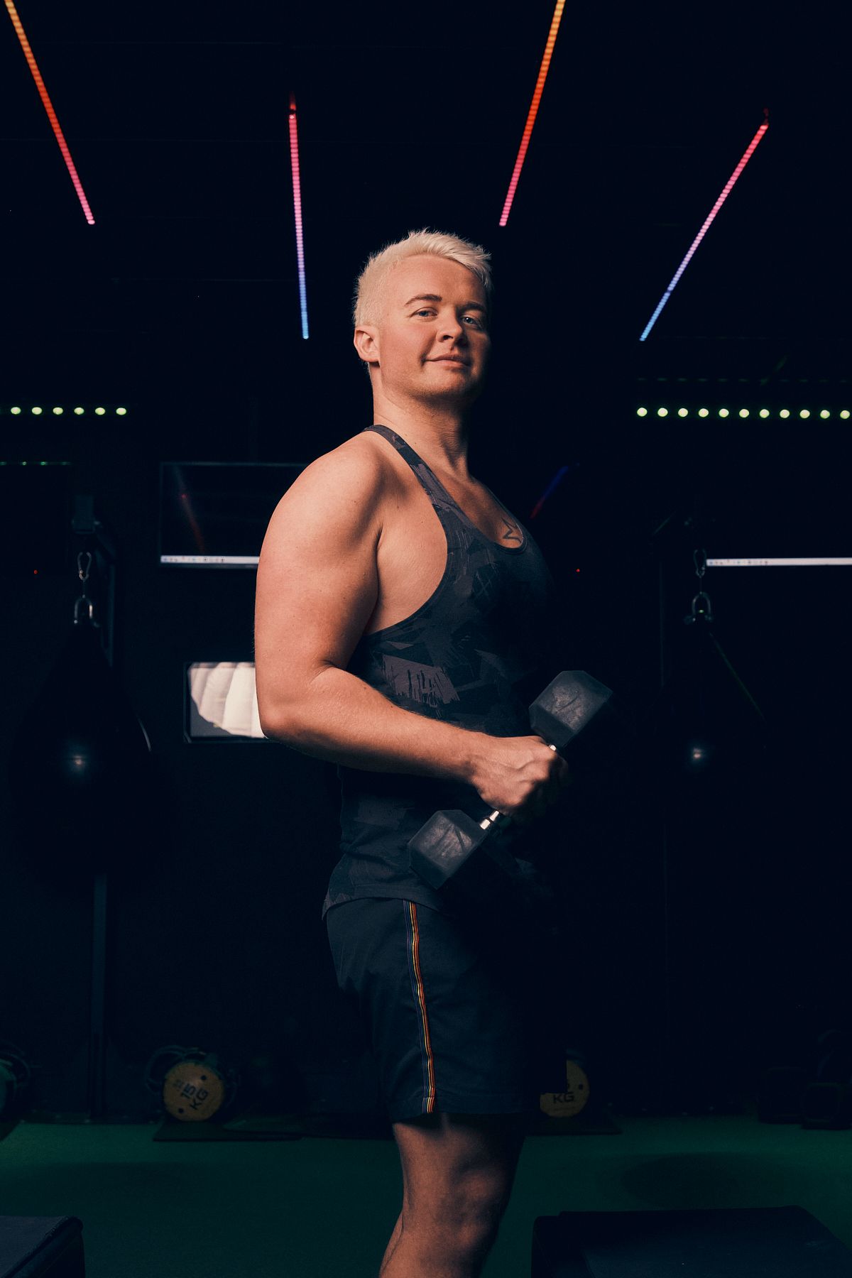 Person with blonde hair in a dark tank top holding dumbbells, posing confidently in a gym with neon lights
