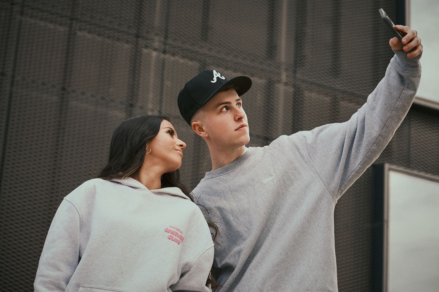 Young man and woman taking a selfie outdoors, both dressed in casual grey sweatshirts with a modern urban backdrop