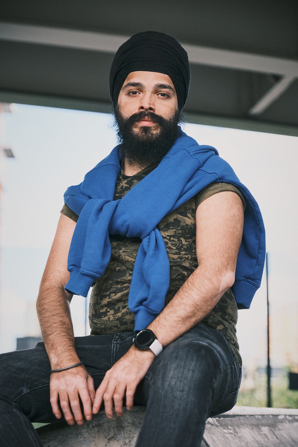 Bearded man in a turban, camo t-shirt, and blue jumper posed confidently, showing personality and style