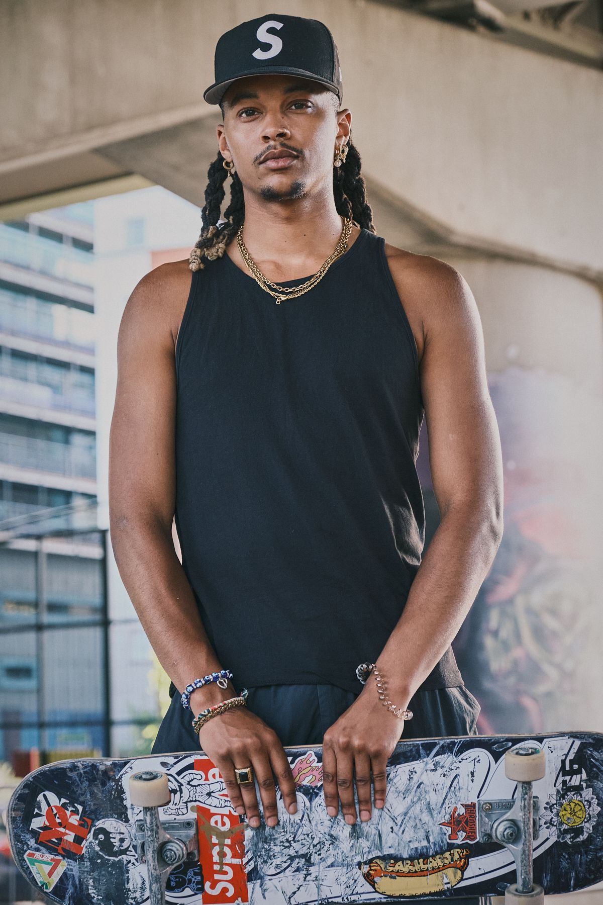 Man in black tank top and cap holding a skateboard, posed under an overpass with a calm expression