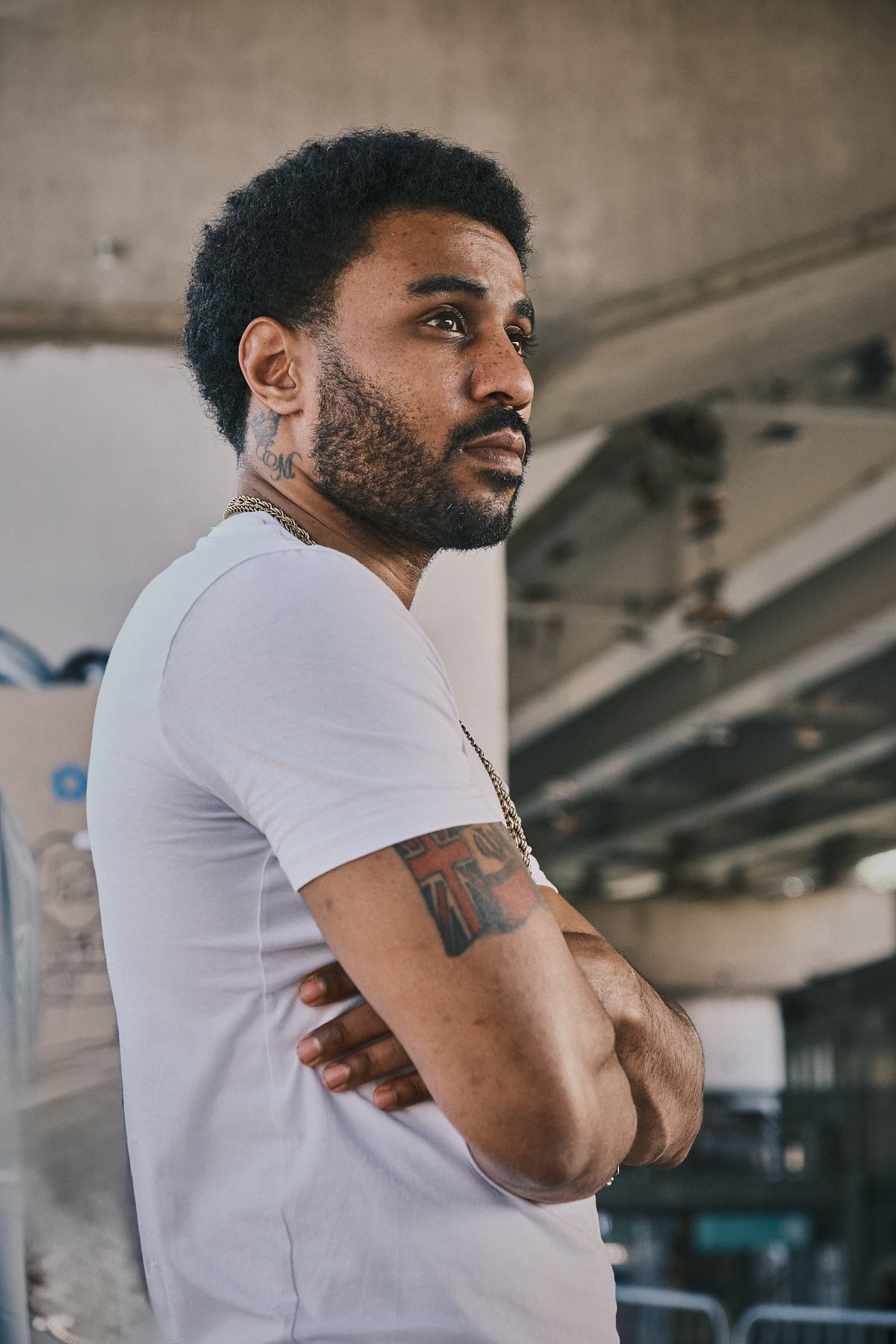 Man in white t-shirt with arm tattoos, standing thoughtfully under a bridge with arms crossed