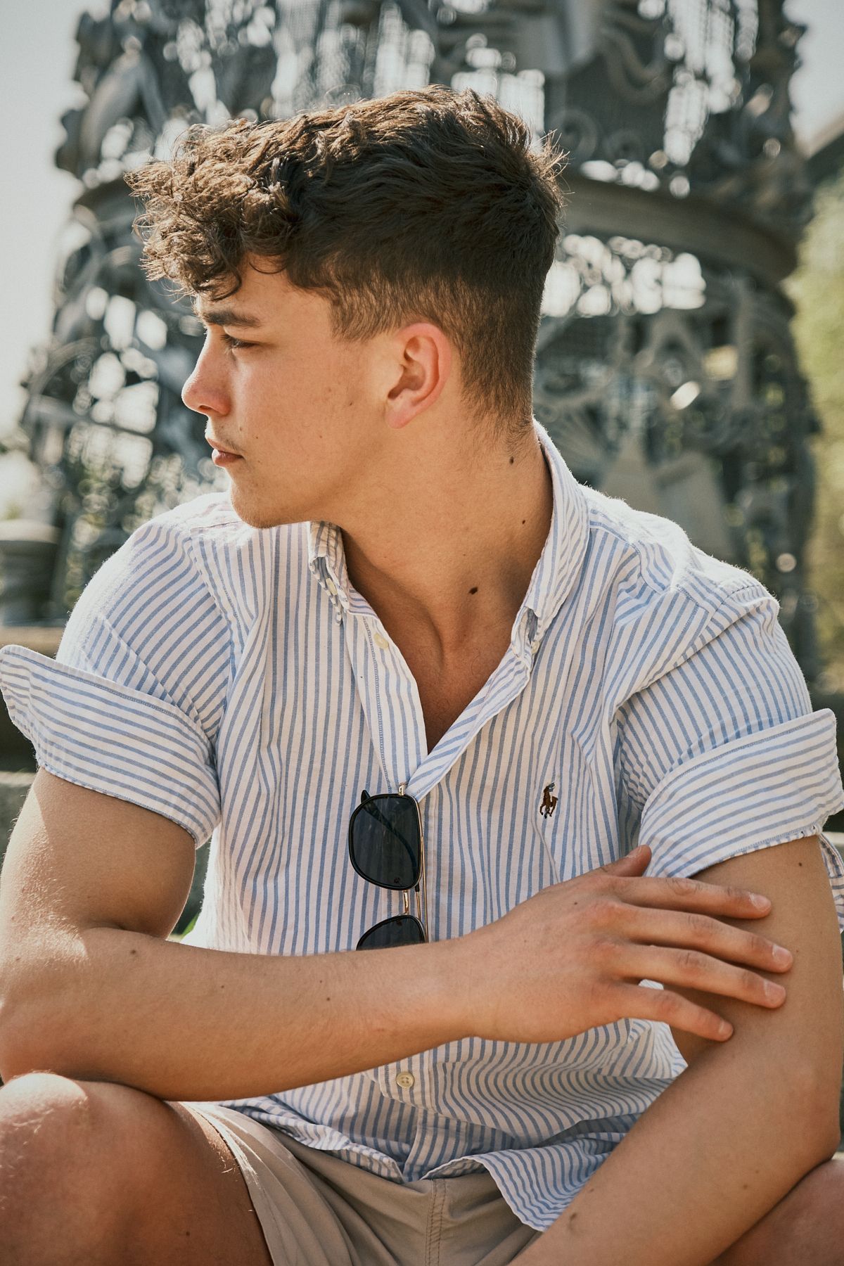 Young man in a striped shirt seated outdoors, looking to the side with a relaxed expression
