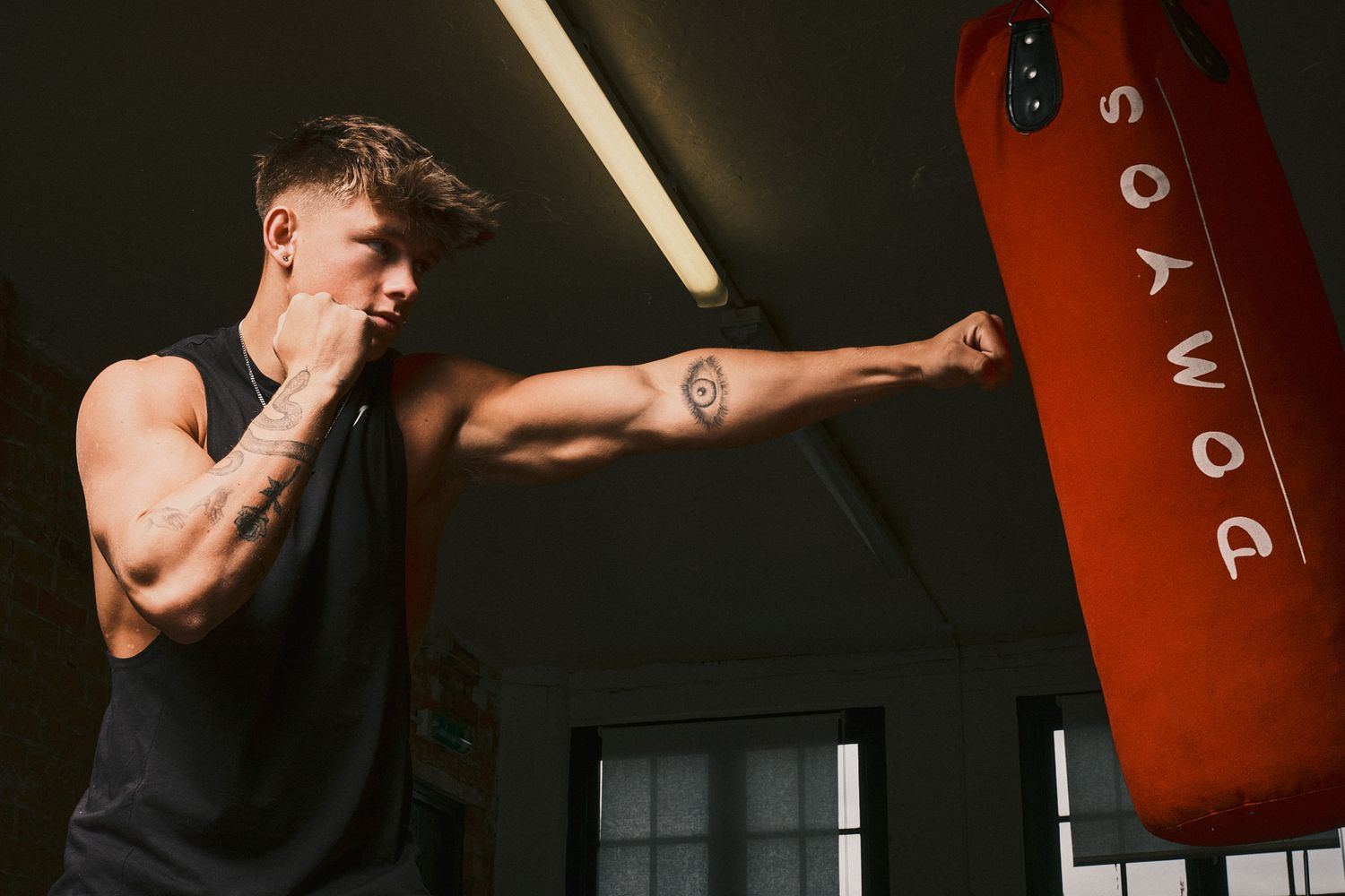 Man in a black tank top throwing a punch towards a red punching bag in a gritty indoor space