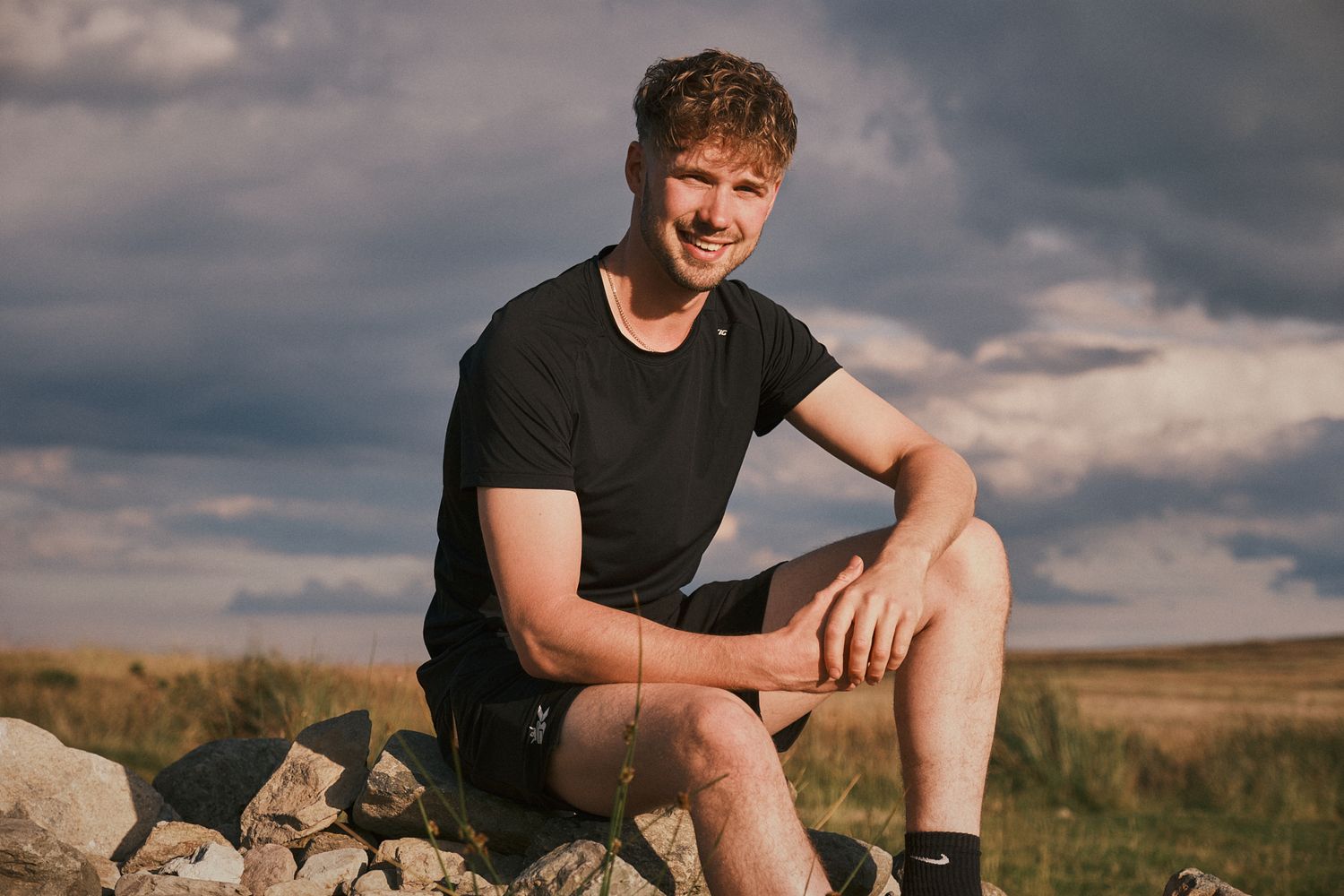 Man in black athletic wear seated on rocks outdoors, smiling in natural sunlight with cloudy sky backdrop