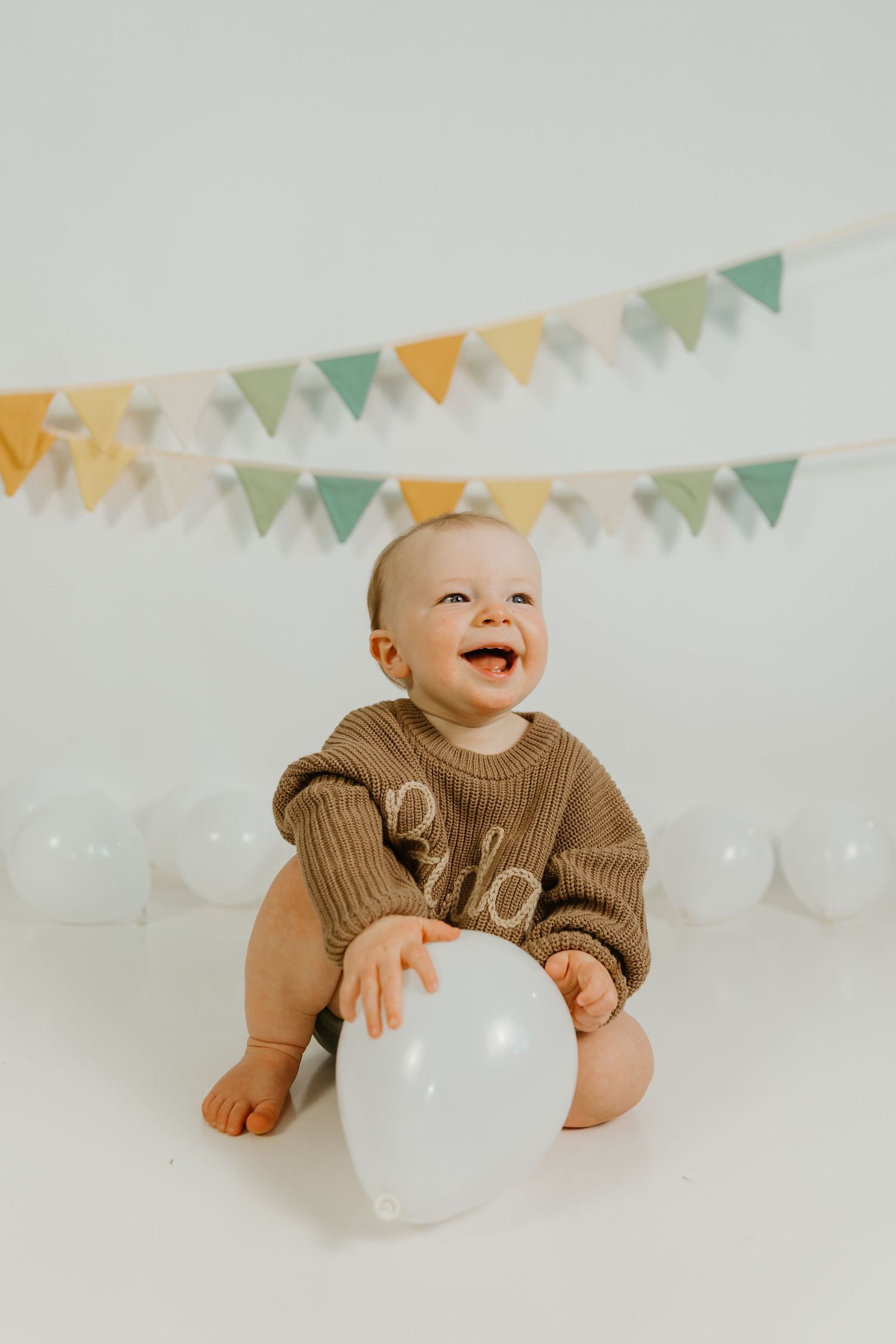 Baby in pink tulle dress sits against rainbow backdrop with pastel balloons and clouds for first birthday.