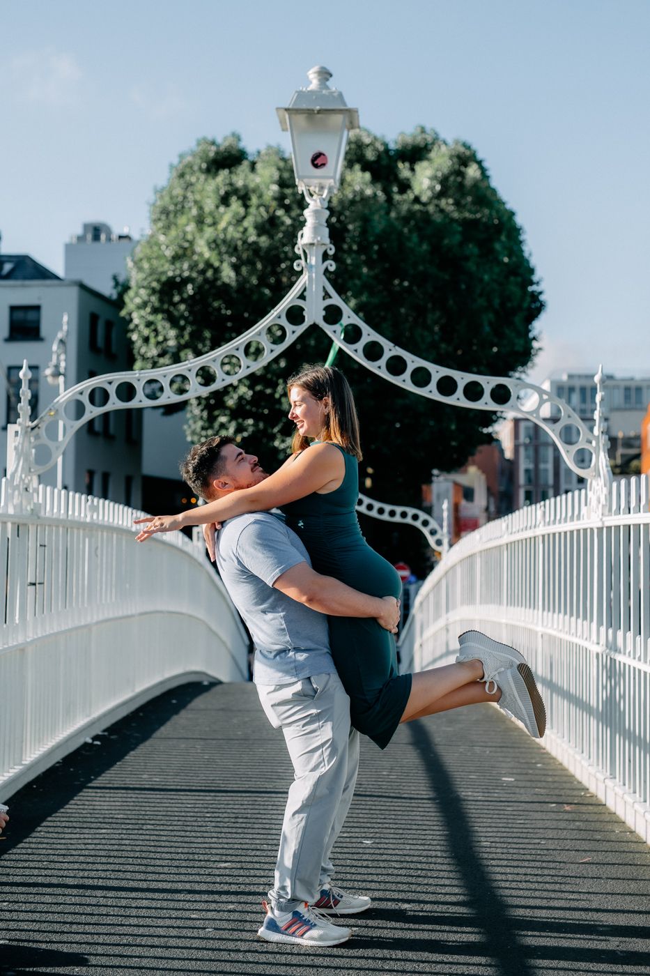 A couple enjoys a romantic moment on a bridge, with one partner lifted joyfully while sunlight bathes the scene.