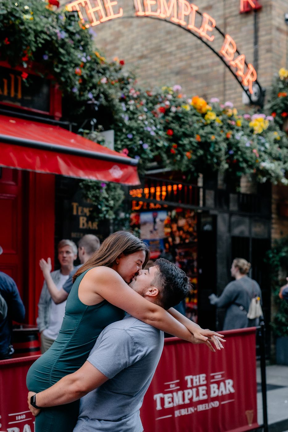 A couple joyfully embraces outside The Temple Bar in Dublin, surrounded by vibrant flowers and a lively atmosphere.