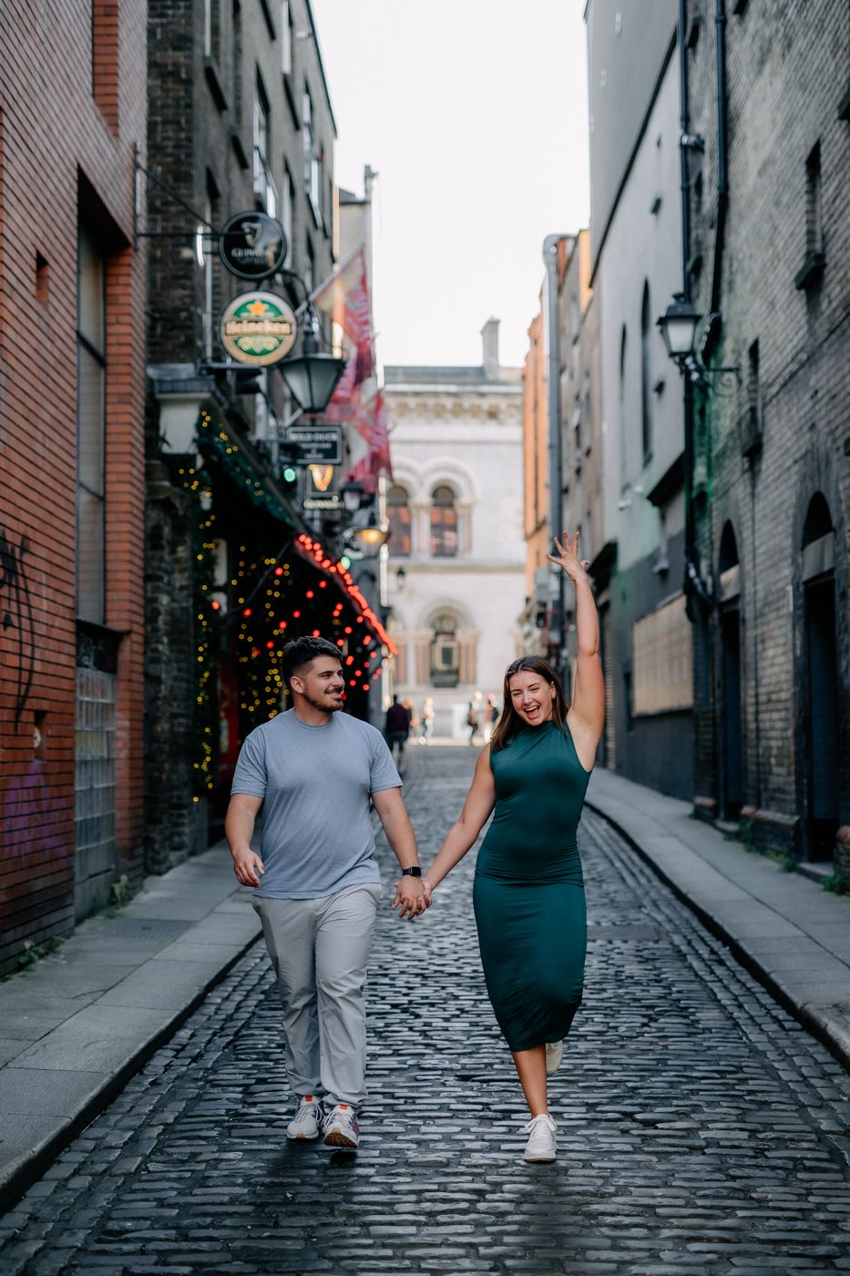 A couple walks hand in hand down a cobblestone street adorned with festive lights, surrounded by historic buildings.