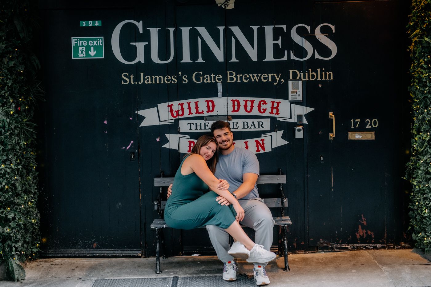 A couple sits closely together on a bench in front of a black door with "Guinness St. James’s Gate Brewery, Dublin".