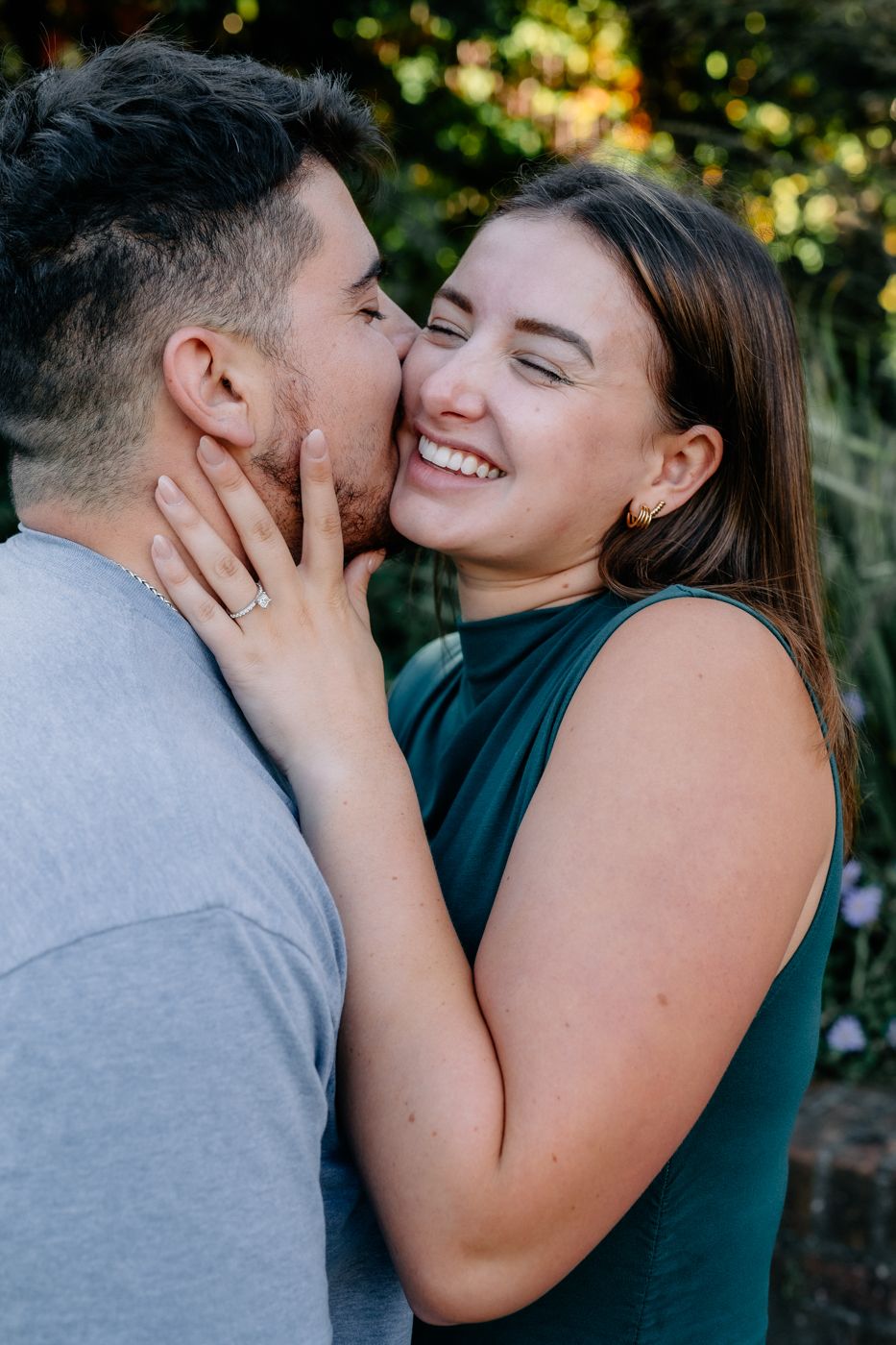 A couple share a tender moment outdoors, with one person gently holding the other's face and displaying a sparkling engagement ring.