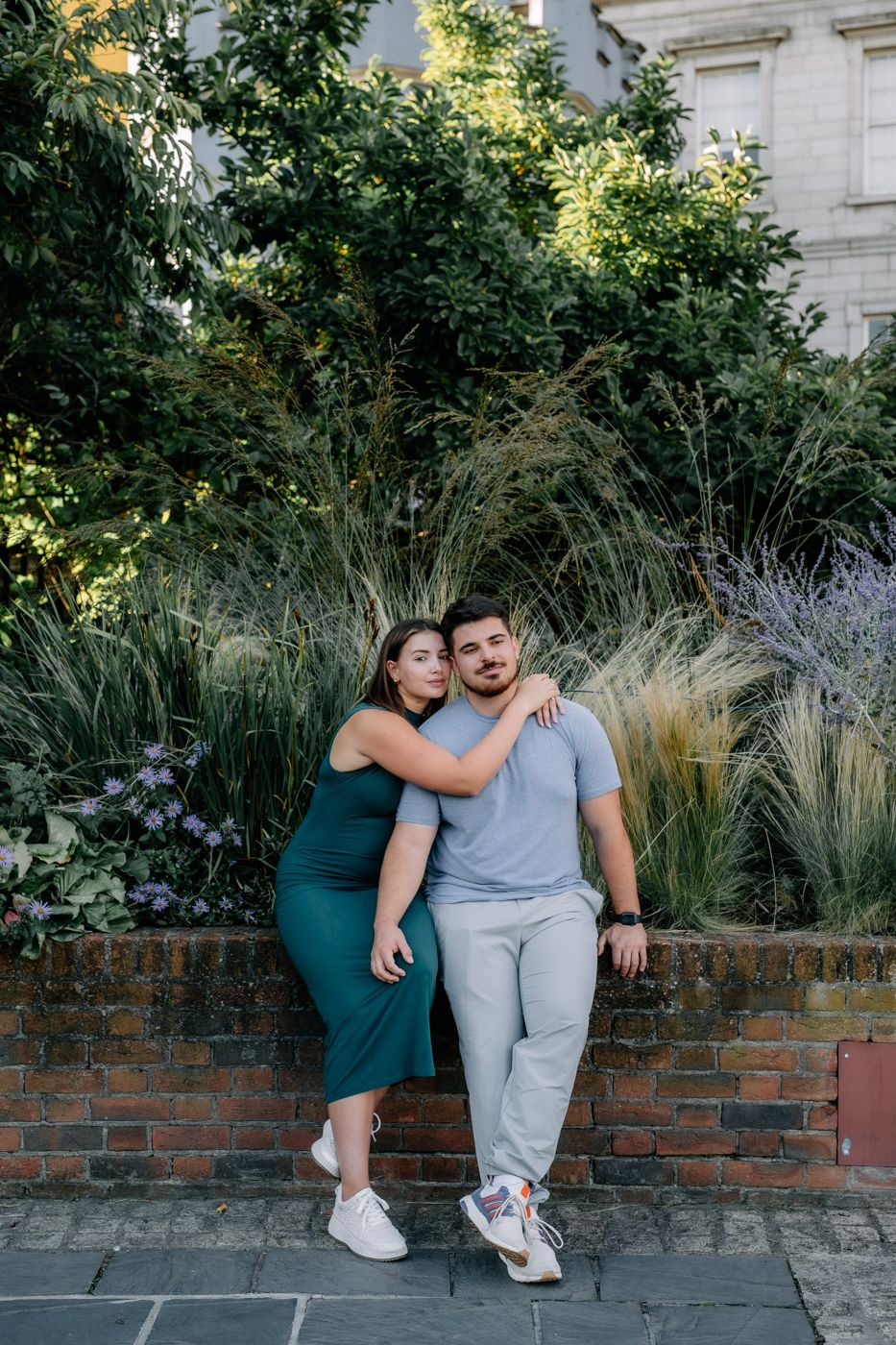 A couple sits closely on a brick wall surrounded by lush greenery and flowers, enjoying a sunny day together.