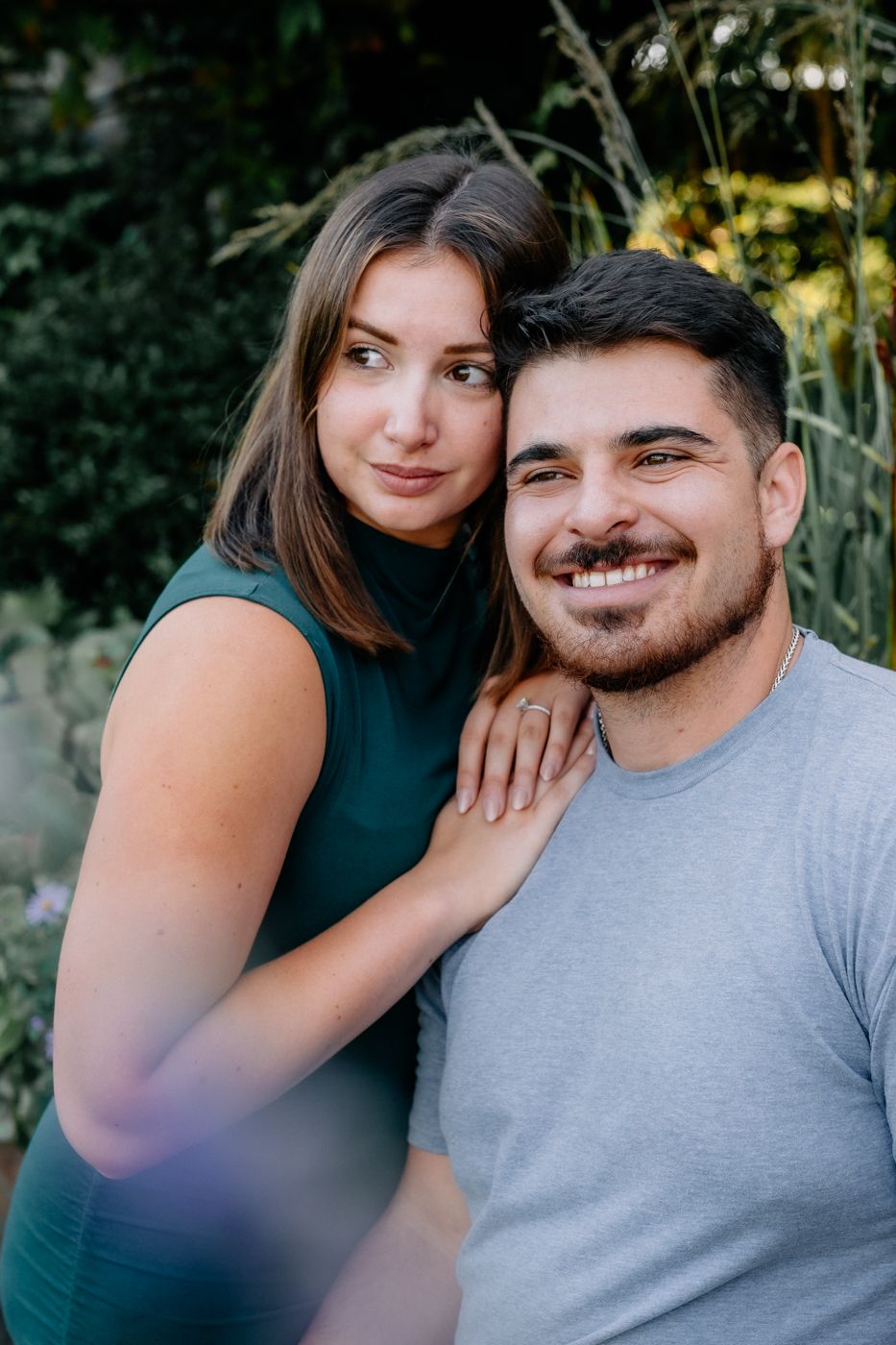 A couple poses closely in a garden, the woman in a green dress with her hand on the man's shoulder, showcasing a loving moment.