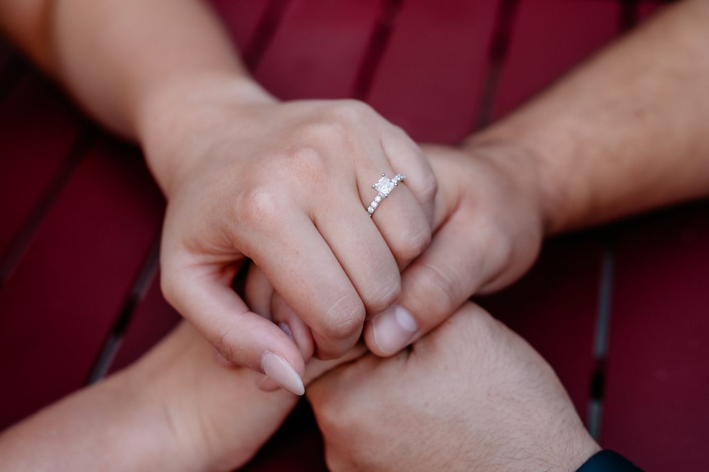 Two hands clasped together, showcasing a sparkling engagement ring on one hand, resting on a red table surface.