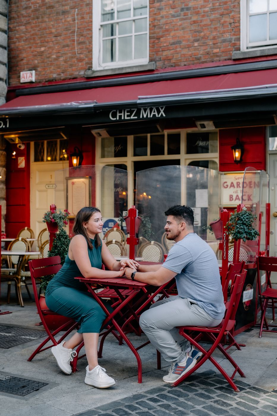 An american couple sits at a red bistro table outside a quaint cafe named "Chez Max," sharing a quiet, intimate moment.