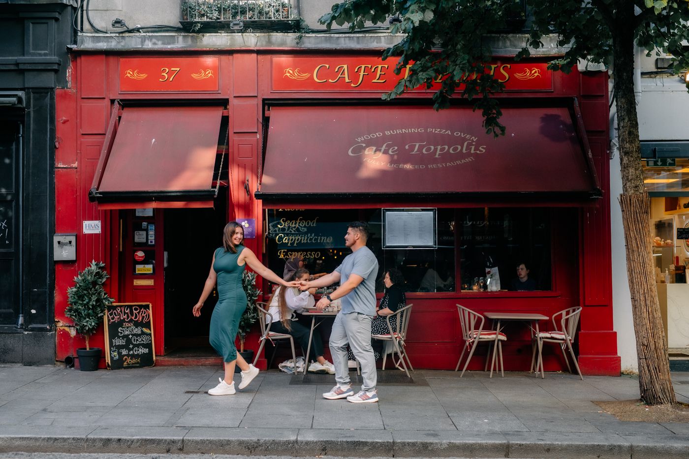 A vibrant red café with outdoor seating, where a couple walks past while another group enjoys coffee indoors on a sunny day. Dublin.