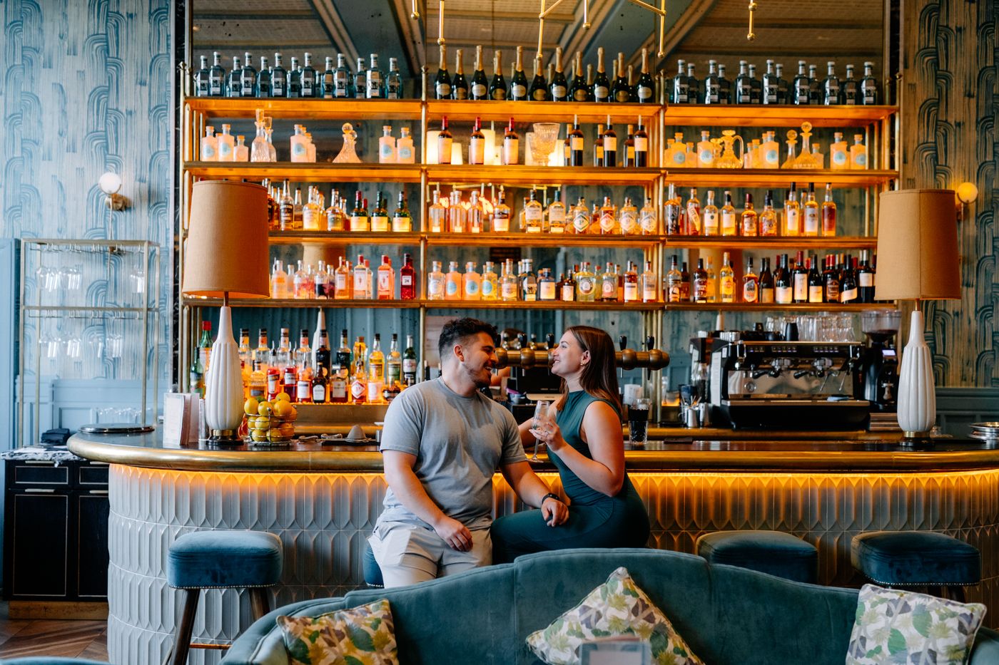 A couple sits at a stylish bar, surrounded by colorful liquor bottles, with one person holding a drink and the other smiling.
