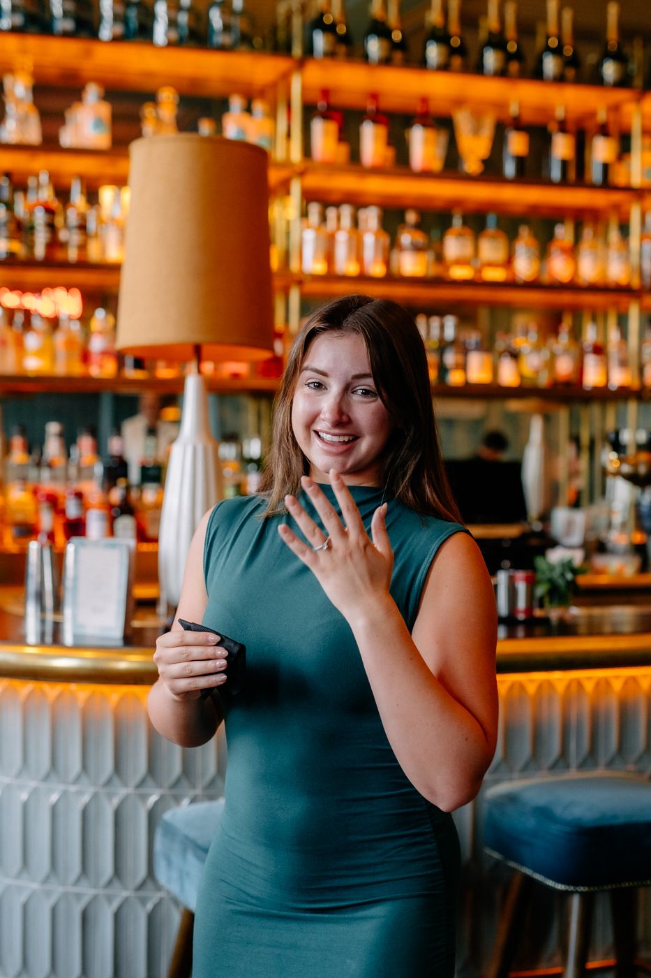 A woman in a green dress holds a phone, standing in front of a well-stocked bar with warm lighting and decorative elements.
