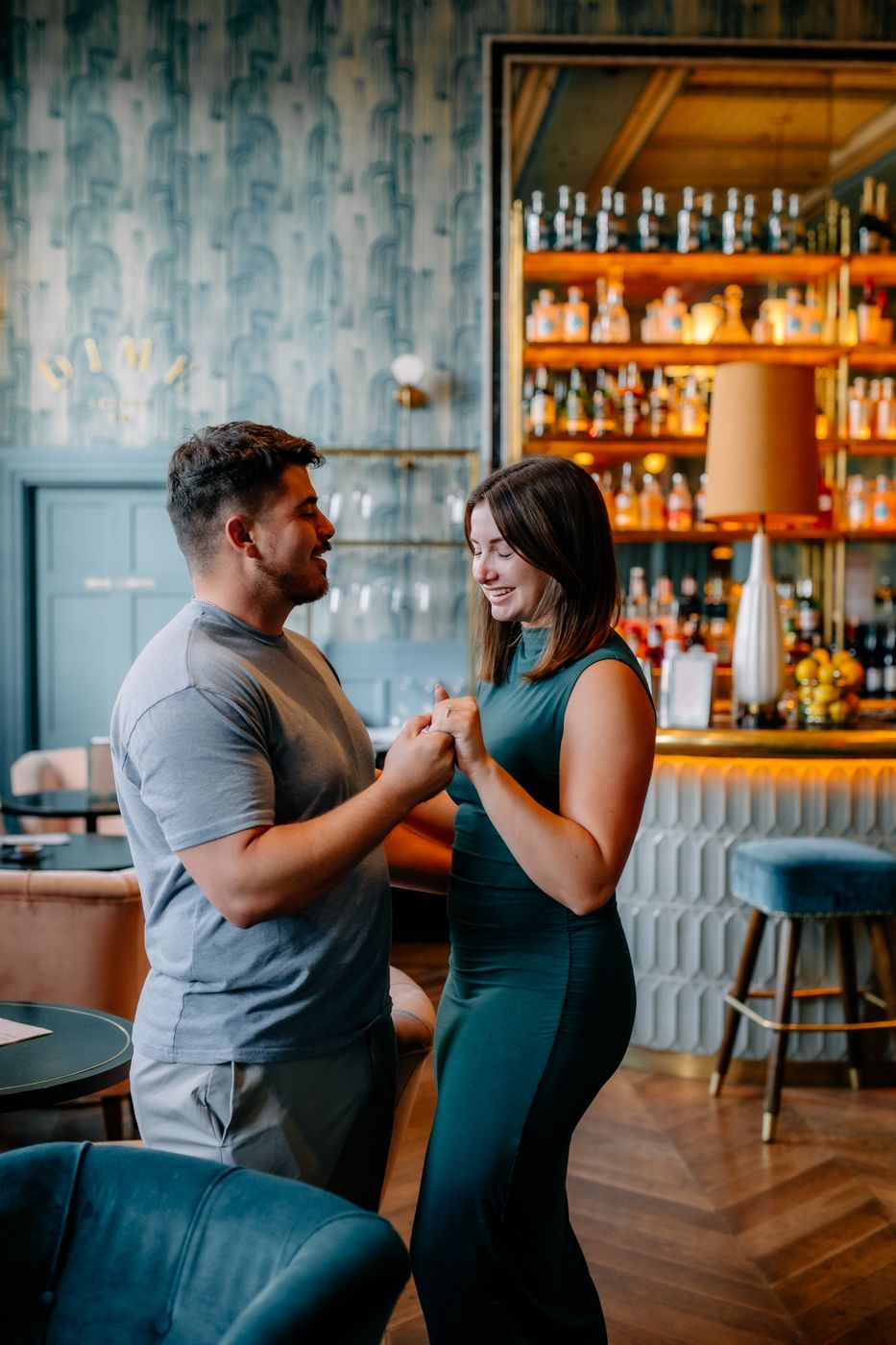 A couple holds hands in a stylish restaurant, surrounded by elegant decor and a glowing bar lined with various drinks.