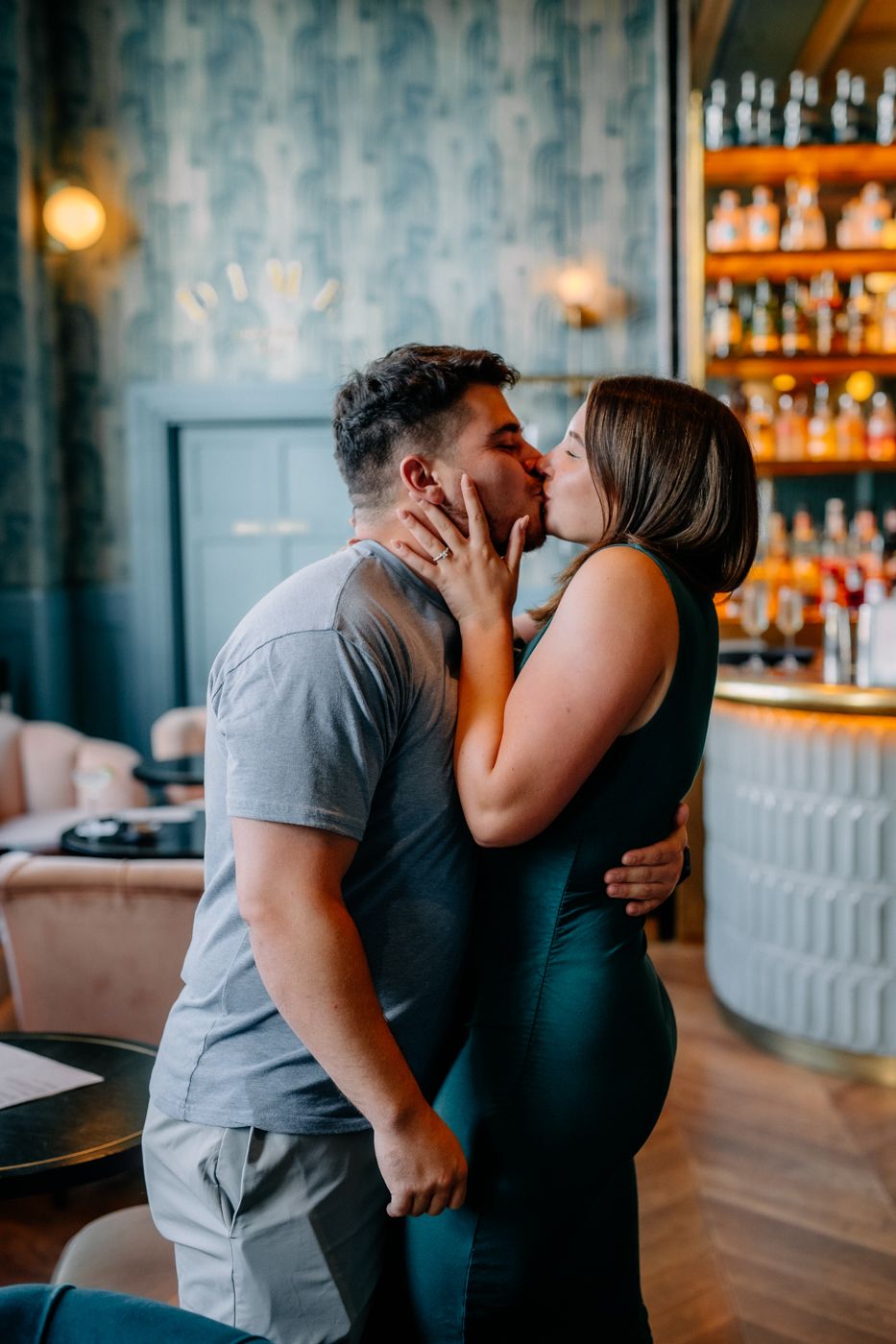 A couple shares a romantic kiss in a stylish restaurant with a cozy ambiance, featuring a well-stocked bar in the background.
