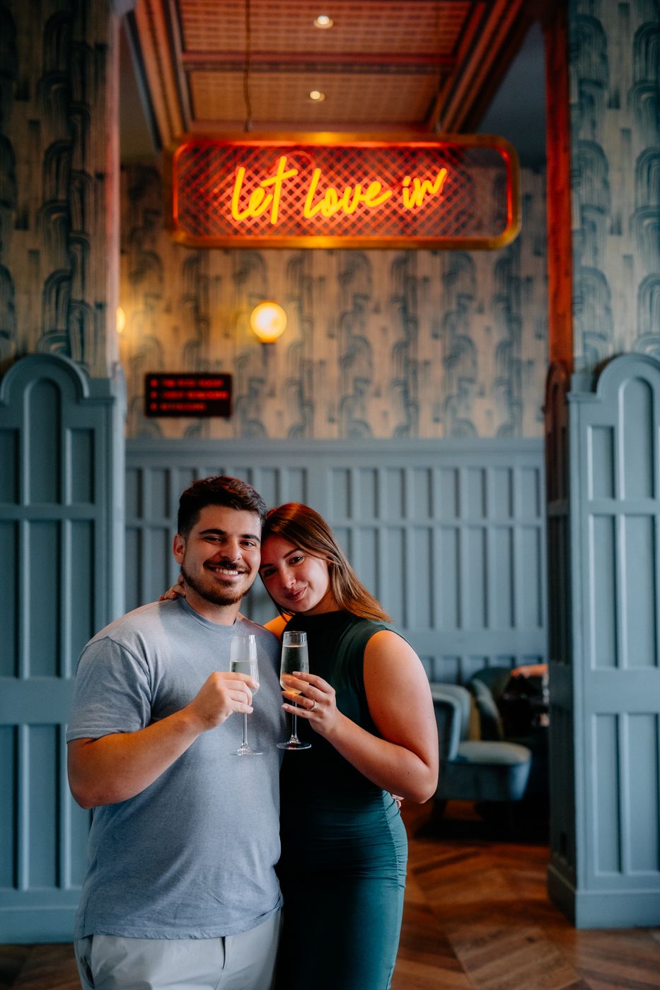 A couple toasts with champagne glasses in a stylish venue, accentuated by a vibrant neon sign reading "let love in."