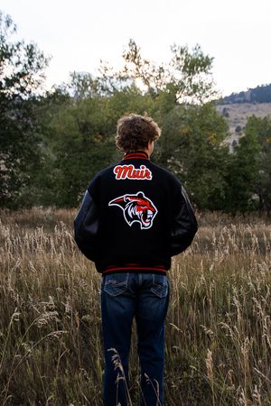 Figure wearing a black sports varsity jacket with a team logo stands in a field of tall grass at sunset.