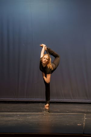 Dancer performs a high leg extension in black attire against a dark backdrop on stage.