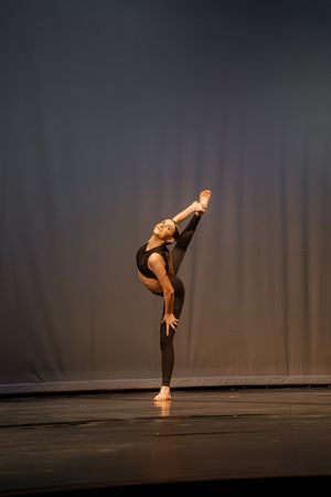 Contemporary dancer performs a dramatic high kick pose on stage in black attire under dramatic lighting.