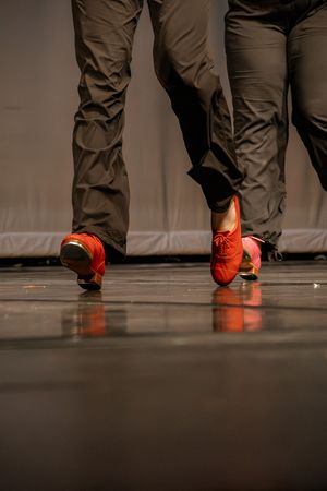 Close-up of red dance shoes and dark pants moving across a stage floor.