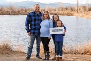 A family of three stands together by a lake on a winter day holding ultrasound photos.