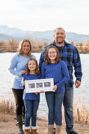 Family wearing coordinating blue outfits poses together by a lake with mountains in the background during sunset.