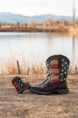 Patriotic cowboy boots decorated with American flag design rest on dirt with a lake view behind them.